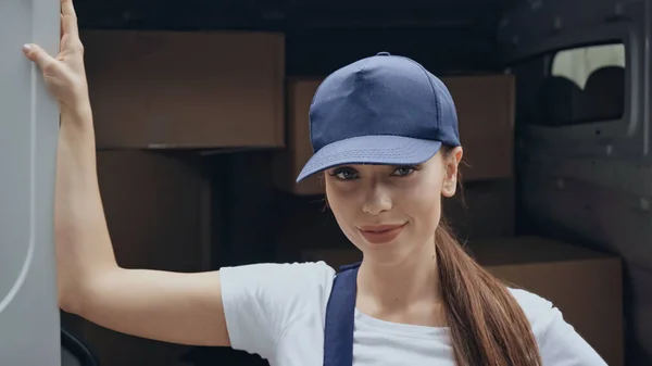 Morena mensajero en la tapa mirando a la cámara cerca de cajas de cartón en el coche al aire libre - foto de stock