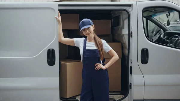 Smiling courier in overalls standing near cardboard boxes in car outdoors — Stock Photo