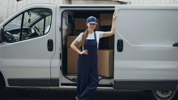 Positive delivery woman looking at camera near boxes in car outdoors — Stock Photo