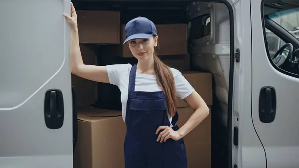 Mujer de entrega en gorra y overoles mirando a la cámara cerca de cajas de cartón en el coche al aire libre - foto de stock
