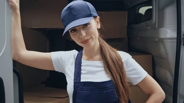 Brunette delivery woman in cap looking at camera near auto outdoors — Stock Photo