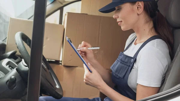 Side view of delivery woman writing on clipboard near boxes in car — Stock Photo