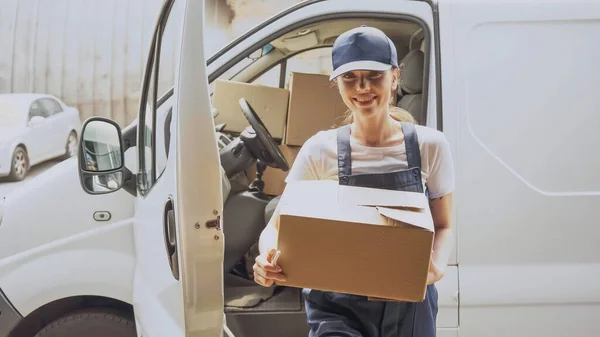 Mujer de entrega sonriente sosteniendo caja de cartón cerca del coche al aire libre - foto de stock