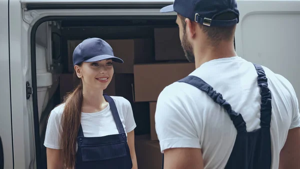 Femme de livraison positive en uniforme regardant son collègue près des boîtes en carton dans la voiture à l'extérieur — Photo de stock