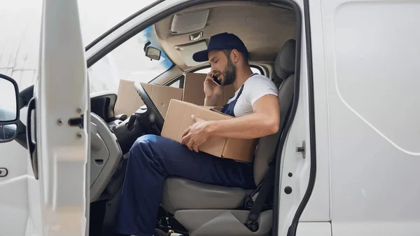 Vista lateral del mensajero hablando en smartphone cerca de cajas de cartón en el coche - foto de stock