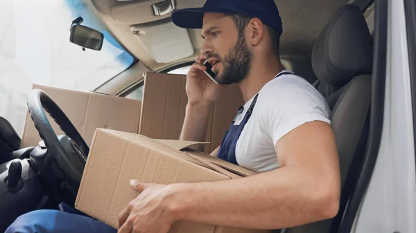 Side view of angry courier talking on smartphone and holding cardboard box in auto — Stock Photo
