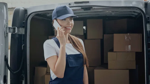 Smiling delivery woman talking on smartphone near cardboard boxes in car outdoors — Stock Photo