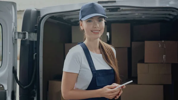 Cheerful delivery woman holding smartphone and looking at camera near blurred boxes in car outdoors — Stock Photo