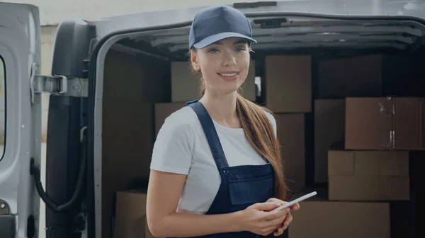 Mujer alegre entrega sosteniendo teléfono inteligente y mirando a la cámara cerca de cajas borrosas en el coche al aire libre - foto de stock