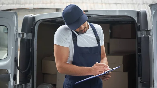 Mensajero en uniforme hablando en el teléfono inteligente y la escritura en el portapapeles cerca de cajas en auto al aire libre - foto de stock