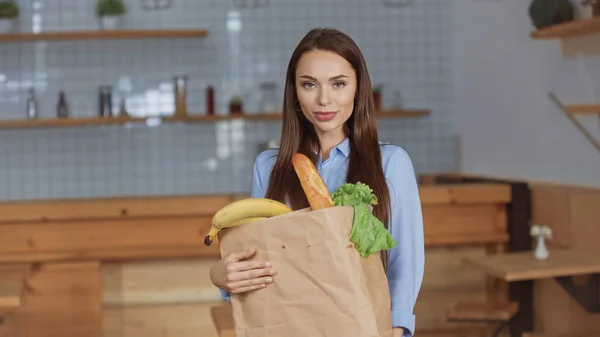 Schöne brünette Frau hält Paket mit Lebensmitteln und schaut in die Kamera zu Hause — Stockfoto