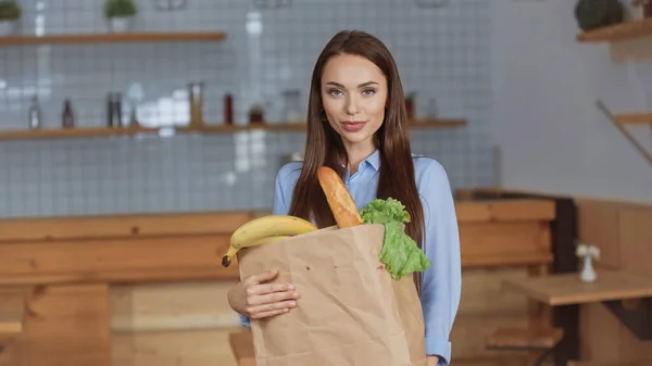 Mujer morena bonita sosteniendo paquete con comida y mirando a la cámara en casa - foto de stock