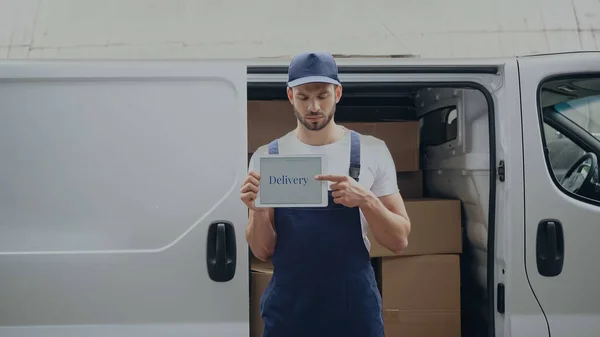 Uomo di consegna in uniforme che punta al tablet digitale con scritte vicino alle scatole in auto all'aperto — Foto stock