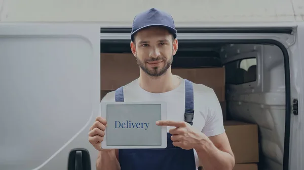 Smiling delivery man pointing with digital tablet with lettering near auto outdoors — Stock Photo