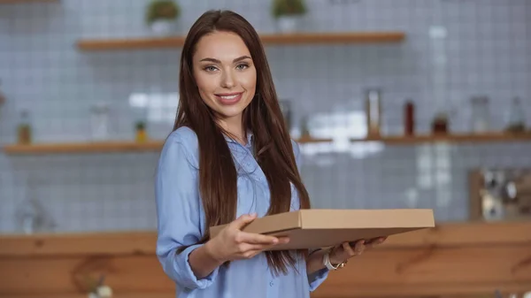 Lächelnde brünette Frau blickt in die Kamera, während sie eine Pizzaschachtel zu Hause hält — Stockfoto