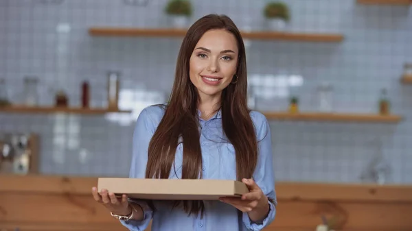 Allegro bruna donna in possesso di scatola di pizza e guardando la fotocamera a casa — Foto stock