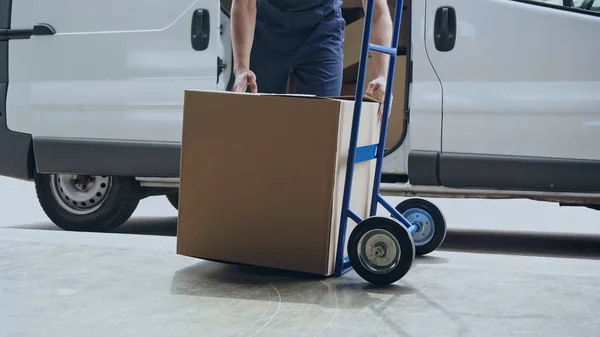 Cropped view of delivery man holding cardboard box near cart and auto outdoors — Stock Photo