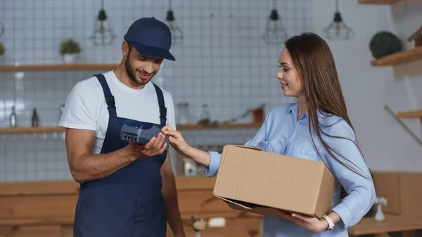 Entrega hombre sosteniendo terminal de pago cerca de la mujer con caja de cartón en casa - foto de stock