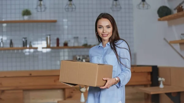 Positive Frau hält Karton in der Hand und blickt zu Hause in die Kamera — Stockfoto