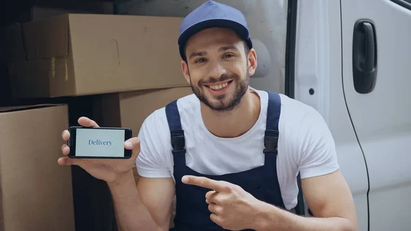 Smiling courier in uniform pointing at cellphone with delivery lettering near carton boxes in car outdoors — Stock Photo