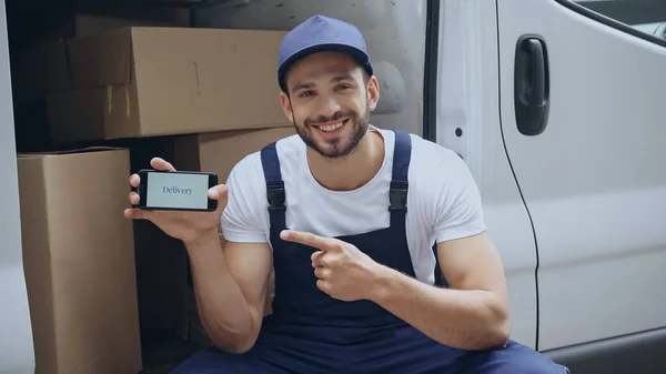 Smiling delivery man in overalls pointing at cellphone with delivery lettering near carton boxes in car outdoors — Stock Photo