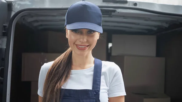 Positive delivery woman in uniform looking at camera near blurred cardboard boxes in car outdoors — Stock Photo