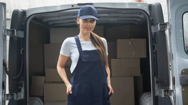 Positivo morena mensajero mirando a la cámara cerca de paquetes en coche al aire libre - foto de stock