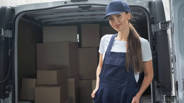 Cheerful delivery woman in cap looking at camera near blurred carton packages in car outdoors — Stock Photo