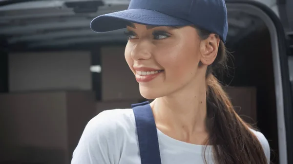 Femme de livraison souriante en uniforme et chapeau regardant loin près des paquets flous en plein air automatique — Photo de stock