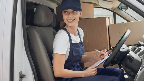 Cheerful delivery woman in uniform writing on clipboard near carton boxes in car — Stock Photo