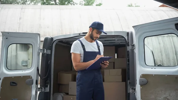 Delivery man in overalls writing on clipboard near cardboard boxes in car outdoors — Stock Photo