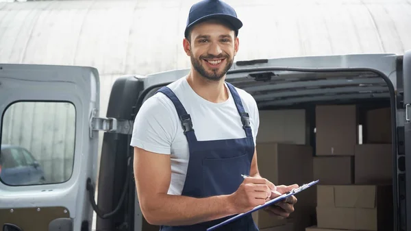 Positive courier in cap writing on clipboard and looking at camera near blurred car with carton boxes outdoors — Stock Photo