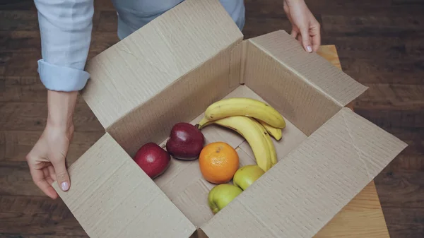 Vista ritagliata della donna che apre scatola di cartone con frutta fresca a casa — Foto stock