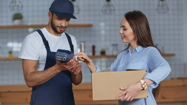 Sorrindo mulher segurando caixa de papelão e usando terminal de pagamento perto de homem de entrega em casa — Fotografia de Stock