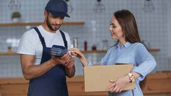 Femme souriante tenant la boîte en carton et utilisant le terminal de paiement près du livreur à la maison — Photo de stock