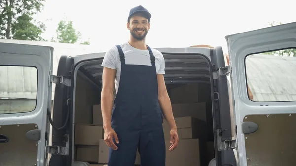 Lächelnder Kurier in Overalls steht neben Päckchen im Auto im Freien — Stockfoto