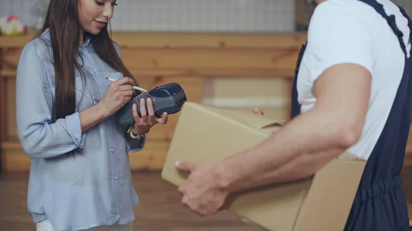 Woman using payment terminal near courier with cardboard box at home — Stock Photo