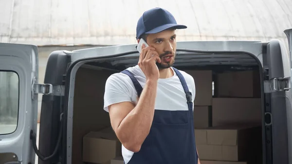 Courier in overalls talking on mobile phone near car on urban street — Stock Photo
