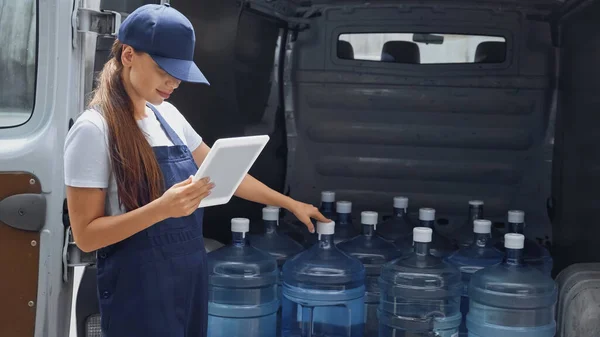 Доставка женщина в комбинезоне держит цифровой планшет возле бутылок с водой в автомобиле на открытом воздухе — стоковое фото