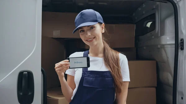 Cheerful delivery woman in overalls holding smartphone with delivery lettering near carton boxes in car outdoors — Stock Photo