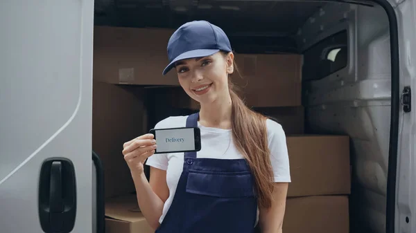 Femme de livraison joyeuse en salopette tenant smartphone avec lettrage de livraison près de boîtes en carton dans la voiture à l'extérieur — Photo de stock