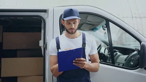 Zusteller schreibt auf Klemmbrett in der Nähe von Kartons im Auto im Freien — Stockfoto