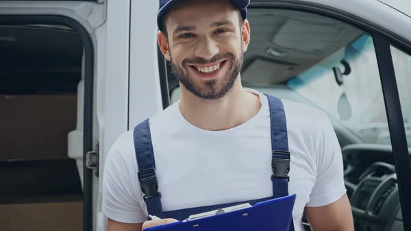 Cheerful courier looking at camera while writing on clipboard near car on urban street — Stock Photo