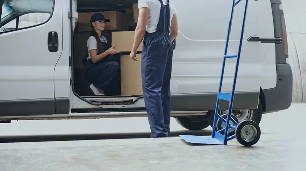 Smiling delivery woman in auto holding carton box near colleague and cart outdoors — Stock Photo