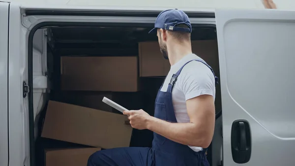 Entrega hombre en overoles sosteniendo tableta digital cerca de cajas de cartón en el coche al aire libre - foto de stock