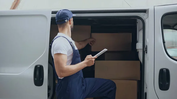 Joven repartidor hombre sosteniendo tableta digital cerca de cajas de cartón en auto al aire libre - foto de stock