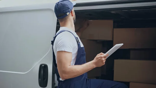 Livreur tenant tablette numérique près des paquets de carton dans la voiture à l'extérieur — Photo de stock