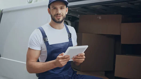 Positive courier with digital tablet looking at camera near cardboard boxes in car outdoors — Stock Photo