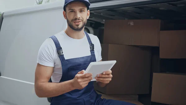 Positiver Kurier mit digitalem Tablet blickt auf Kamera in der Nähe von Kartons im Auto im Freien — Stockfoto