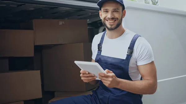 Cheerful courier holding digital tablet near carton boxes in car on urban street — Stock Photo